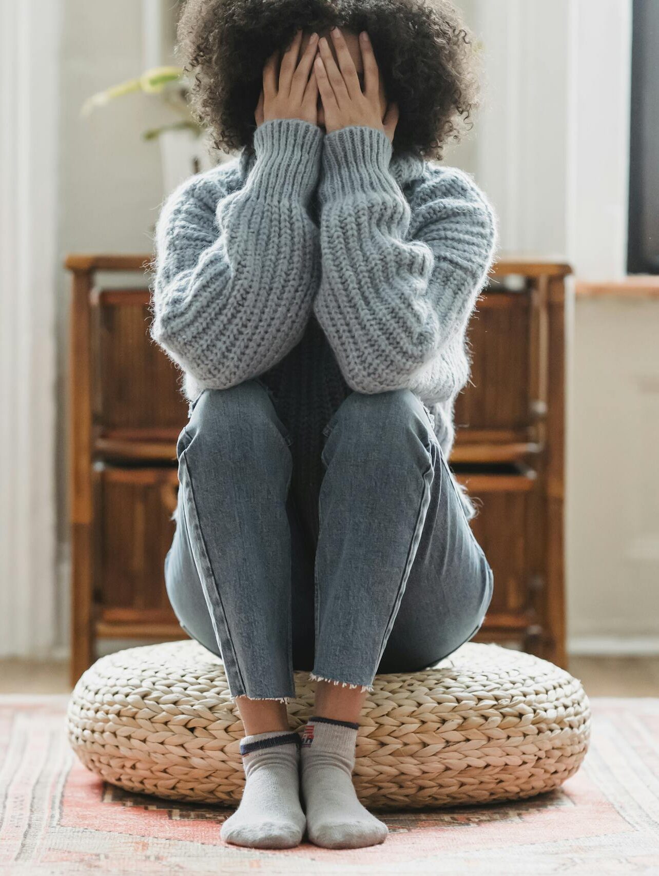 Full body of anonymous depressed female in casual clothes sitting on rattan stool in light room with windows at home