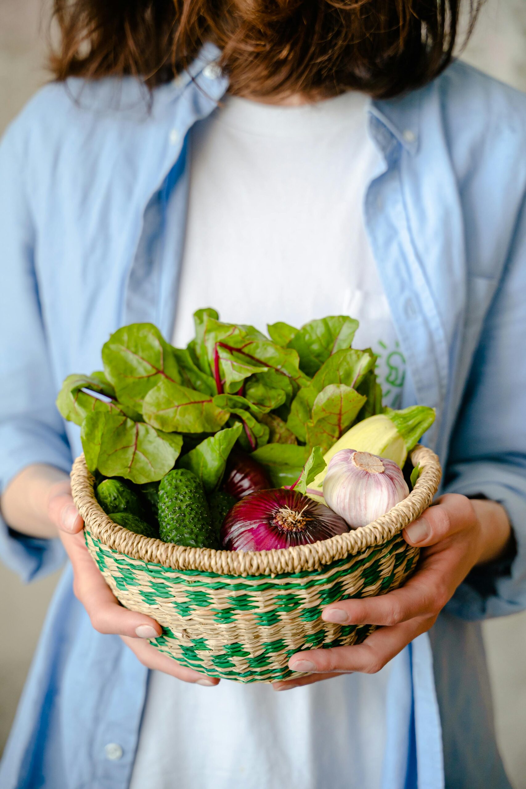 woman holding a woven basket of fresh vegetables, wearing a blue button up shirt and a white t-shirt